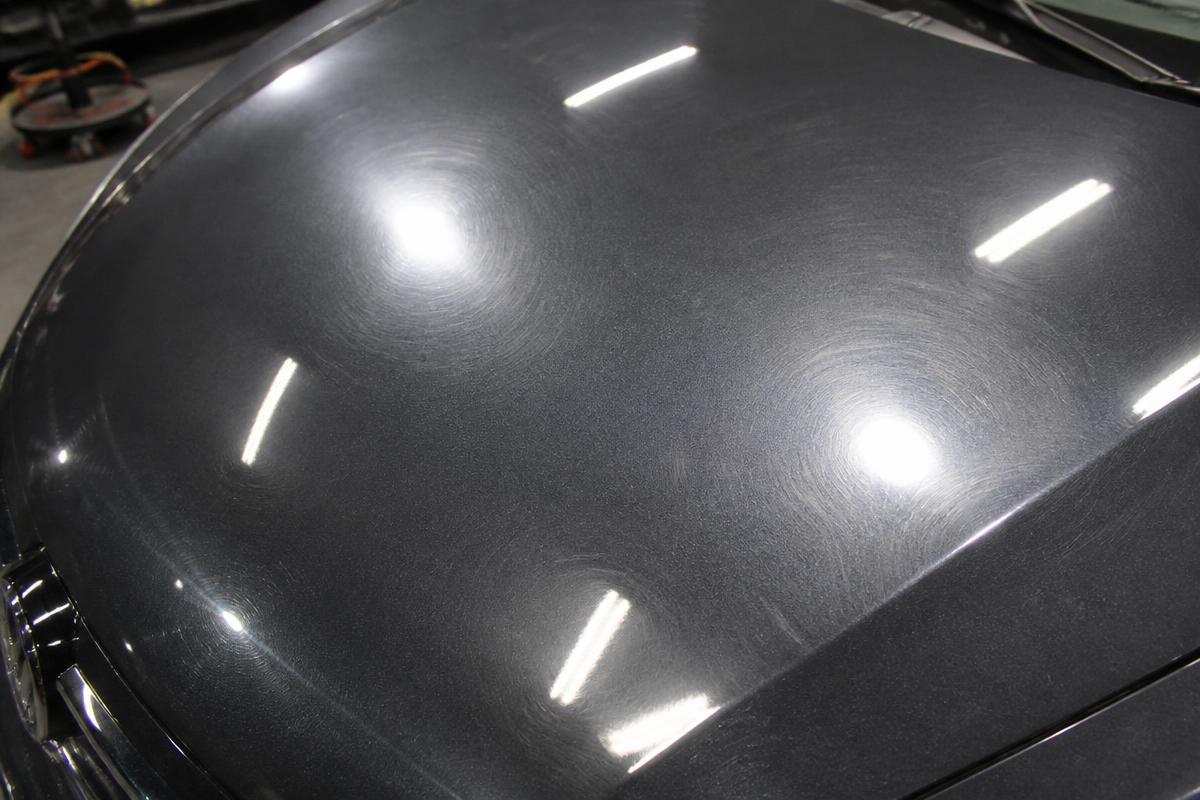 Dark metallic car hood showing heavy swirl marks under inspection lights before paint correction